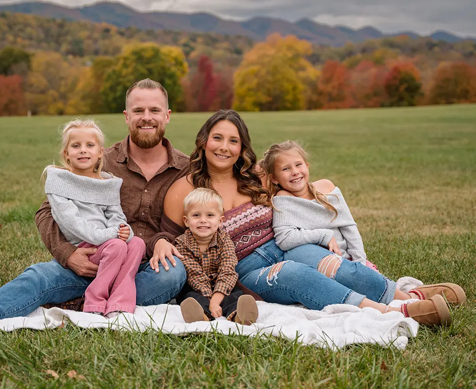A family of five smiling together on a blanket in a scenic outdoor field with autumn trees.